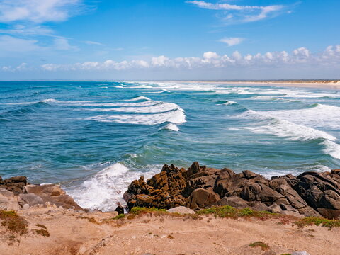 Quimper, France - 04.07.2015: Dynamic white waves rolling onto a rocky coastline with a distant sandy beach under a bright blue summer sky