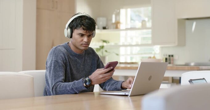 Man in sweater with headset after alert, scrolling pink phone and tapping laptop in kitchen smiling