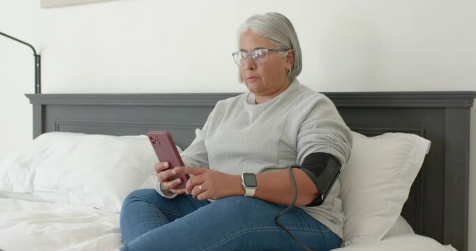 Mature African American woman sitting on bed, looking and tapping pink phone while checking BP cuff