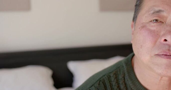 Senior Asian man sitting on bed in green sweater, camera reframing to reveal shoulder, copy space
