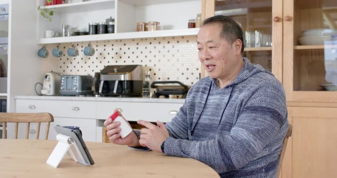 Asian mature man sitting facing tablet holding white bottle red cap, opening and showing contents