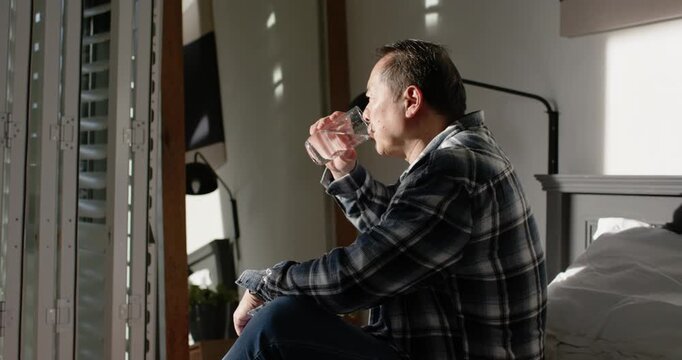 Asian man sitting on bed holding clear glass, drinking water while observing shutter light patterns
