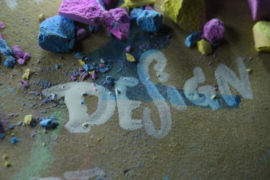 A close-up studio shot of the word "DESIGN" handwritten on a brown textured background, decorated with bright blue, pink, and yellow broken chalk pieces and powder.