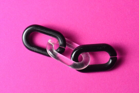 A top-down studio shot of three interconnected plastic chain links, featuring two black links joined by a transparent clear link in the middle. Isolated on a vibrant pink background.