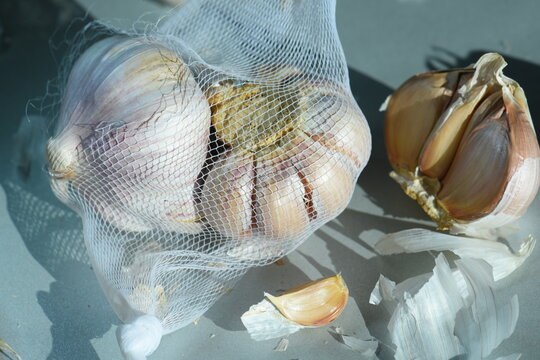 Top view of whole garlic bulbs packaged in a white mesh bag beside an opened garlic head with dry husks on a gray surface under hard daylight.
