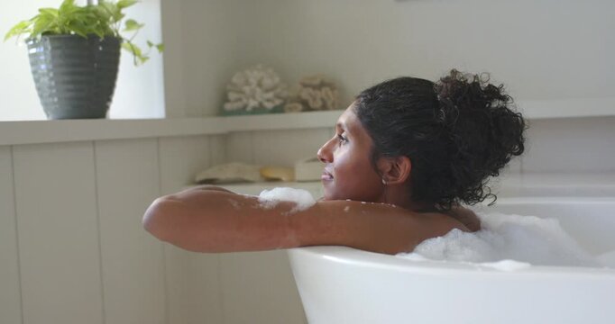 African American woman reclining in white bathtub, tracing foam, following window light, breathing