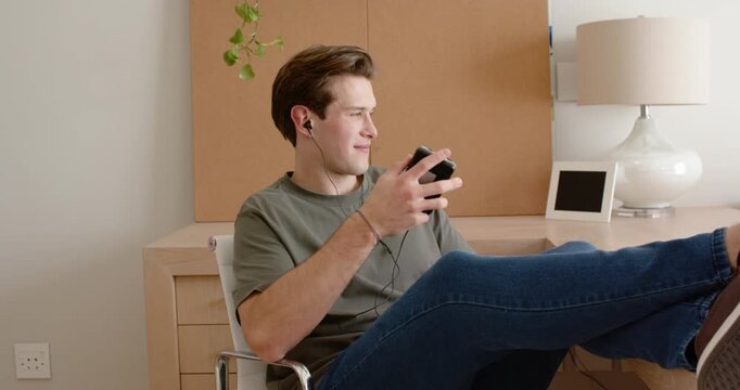 Man mid-20s reclining in metal chair with feet on desk, using black device, responding to sound