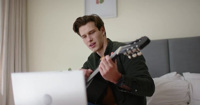 Man in mid-20s placing guitar, following laptop, adjusting fingering and strumming on bed