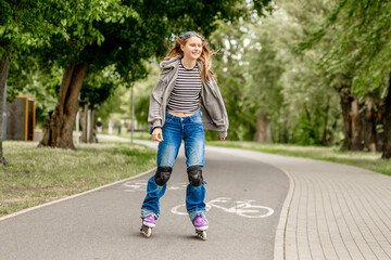 Naklejka premium Happy young girl rollerblades on paved park path. Green trees line route. Sunny daytime activity.