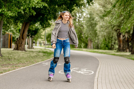 Happy young girl rollerblades on paved park path. Green trees line route. Sunny daytime activity.