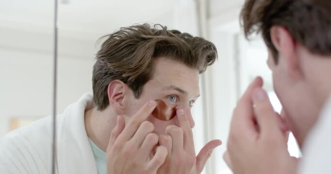 Adult male starting under-eye skincare, leaning into mirror in bathroom, applying copper patch