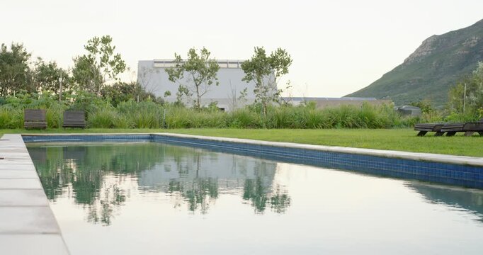 Camera is starting to pan right to reveal long pool reflecting trees and rippling, showing loungers
