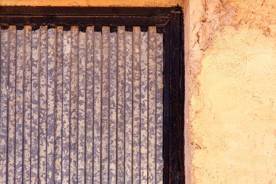 A close view shows a rectangular vertical corrugated galvanized metal panel inside a dark frame. Beside the metal panel is a rough textured yellowish orange earthen wall showing uneven surfaces and cr