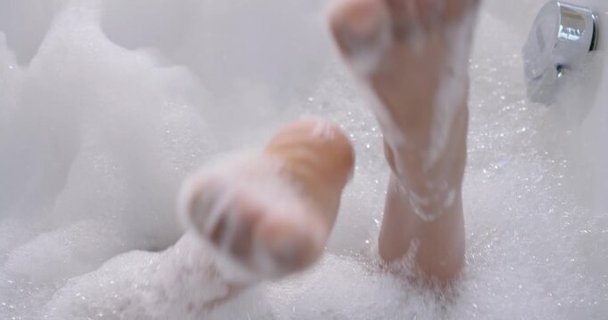 Leaning back, adult woman is reclining in freestanding tub, raising legs through foam to relax