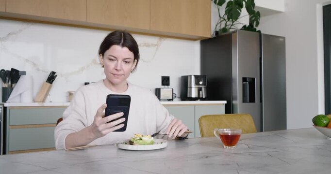 Woman sitting at kitchen island, spotting smartphone, reaching for it, reading funny text, smiling