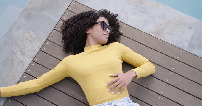 Adult African American woman in yellow top lying on deck by pool, sun urging adjusting shades