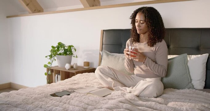 African woman in beige top sitting in loft journaling in notebook then reaching tablet holding mug