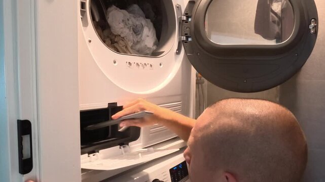 Man performing routine maintenance of a household tumble dryer, removing lint and dust from the filter compartment using a vacuum cleaner, cleaning brush and cloth. Domestic appliance cleaning and hom