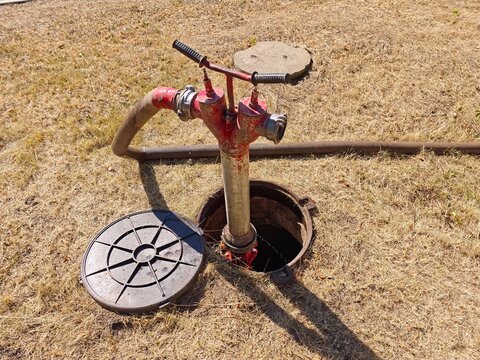 Close up of an underground fire hydrant standpipe with an attached hose connected through an open manhole on dry grass for municipal water extraction.