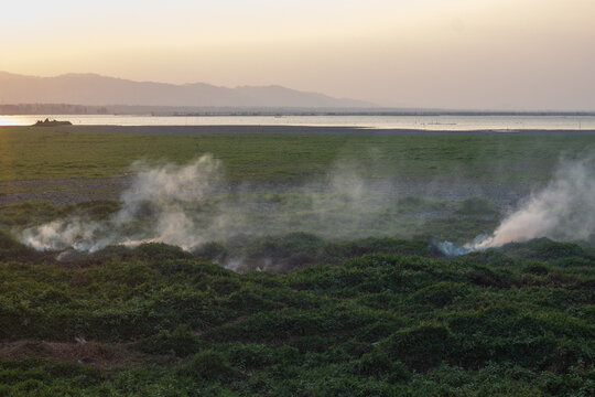 Smoke Rising From Grassland Near Lake at Sunset