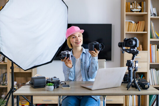 A smiling caucasian woman sits at a desk with multiple cameras and photography equipment, ready to create content