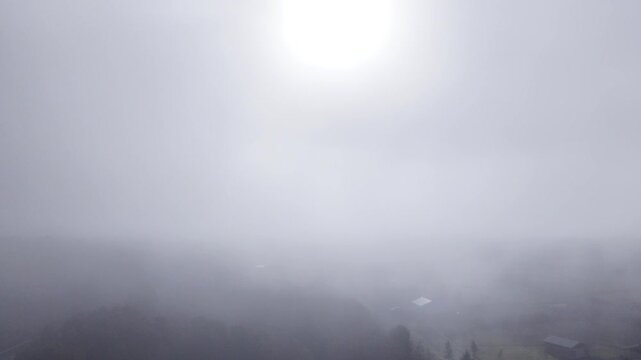 Drone footage rises into a heavily misty rural landscape as the scenery quickly disappears beneath a dense wall of fog, leaving only the faint glow of the rising sun visible through the mist.