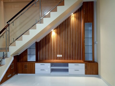 Modern staircase with glass railings and wooden paneling. White cabinets and drawers below offer storage and style. Warm lighting highlights the vertical wood grain design