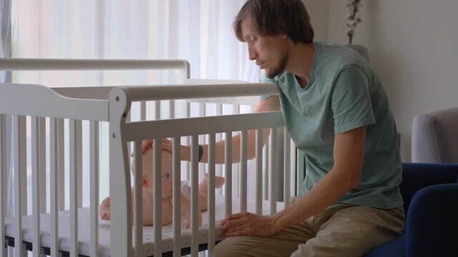 Sleepy father stroking five-month-old son in crib while falling asleep. Tender family moment.