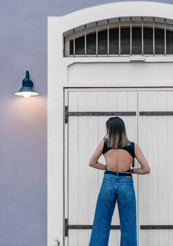 Back view of a young woman in a backless black top and blue jeans standing in front of white wooden doors near a burning wall lamp.