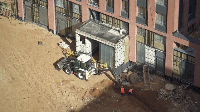 Heavy machinery excavating a deep pit on a construction site, while a worker in overalls stands by and supervises the earthmoving operation during a sunny day. Powerful excavator bucket extracts groun