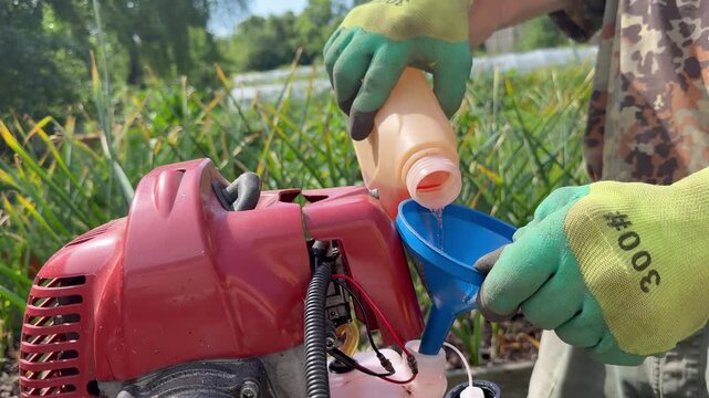 Person pouring fuel into a brush cutter engine tank with a funnel, lawn care preparation, yard work, gardening tools