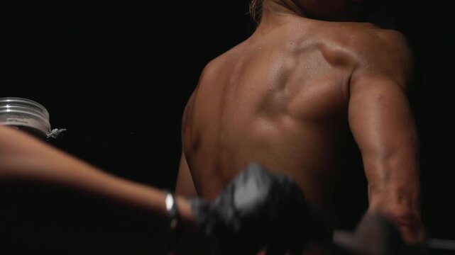 Close-up of technician applying spray tan to woman's lower back and upper thigh in studio under warm directional light, skin shimmering with fresh tanning solution