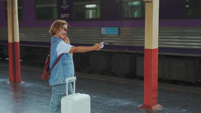 A cheerful mature woman traveler in denim stands on a train platform with her white suitcase and takes a selfie while waving joyfully at the camera as a purple train waits behind her capturing the exc