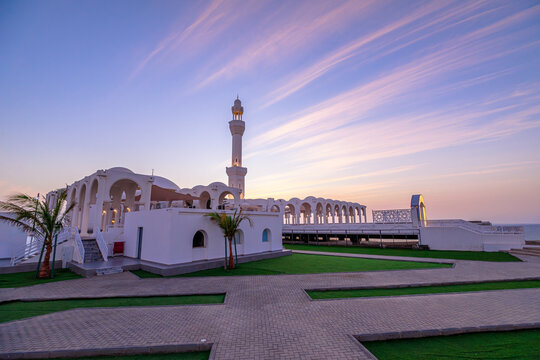 Al Rahmah Mosque, a striking white building with a tall minaret, standing on Jeddah Corniche and appearing to float above the Red Sea as sunset casts vibrant colors across the sky in Saudi Arabia