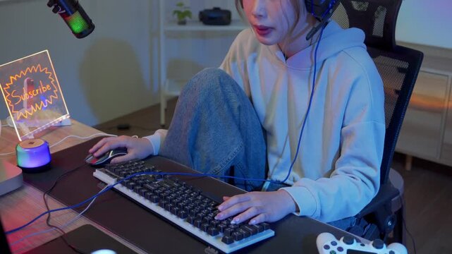 Young woman gamer streaming live online at night playing on a computer with headset and RGB keyboard in a colorful neon lit home setup with microphone and subscribe sign