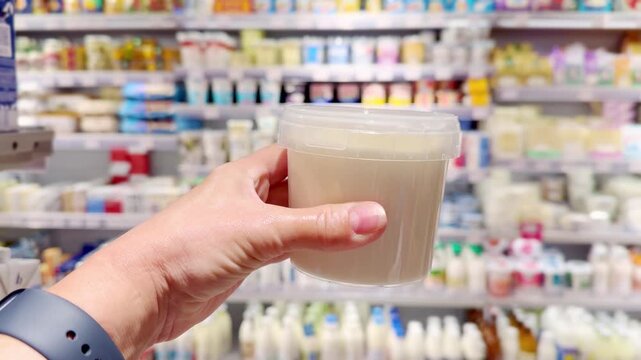 Hand Holding Yogurt and Cottage Cheese Packages in Supermarket Dairy Section &ndash; Dairy Products Against Shelves Background