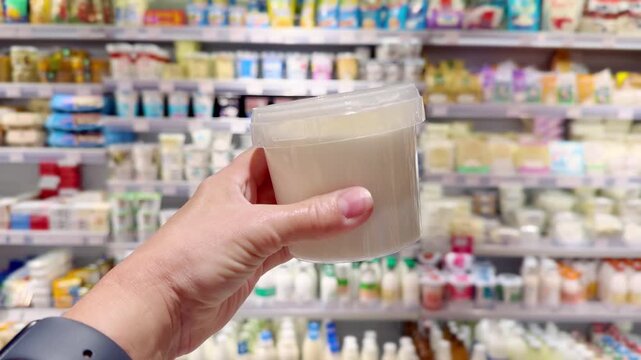 Hand Holding Yogurt and Cottage Cheese Packages in Supermarket Dairy Section &ndash; Dairy Products Against Shelves Background
