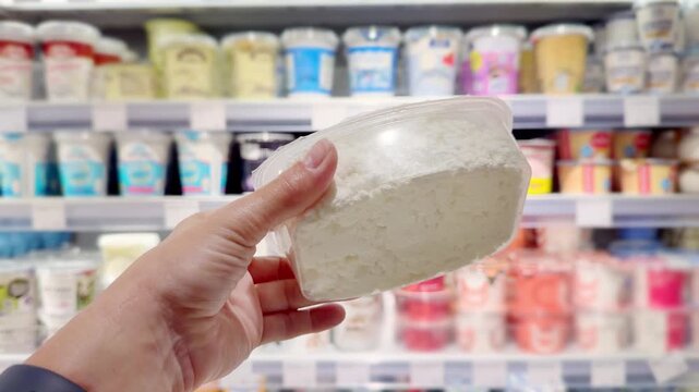 Hand Holding Yogurt and Cottage Cheese Packages in Supermarket Dairy Section &ndash; Dairy Products Against Shelves Background