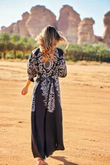 Young woman wearing long dark dress, walking on desert landscape, palm trees and large rock formation typical for Al Ula region in distance. View from behind © Lubo Ivanko