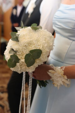The bride holds a lush white wedding bouquet of small flowers and green foliage, decorated with long strands of pearls.