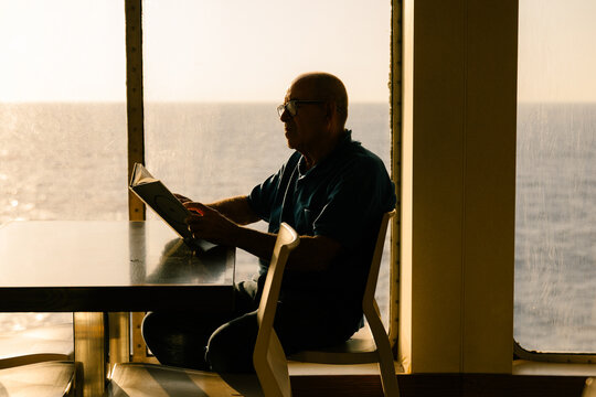 Elderly man reading a book by a ship window in the sunset light.