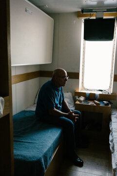 Wide shot of elderly man relaxing as he sits on the bed in his cabin