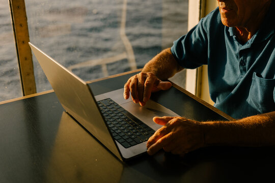 Elderly man working on a laptop by a window overlooking the ocean.