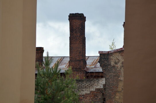 View of an old weathered red brick chimney rising from a rusty tin roof of a ruined industrial building. Abandoned factory architecture with a green pine tree under an overcast sky.