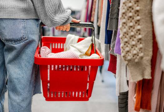 Customer holds red basket with soft garments while walking past clothing racks in shop