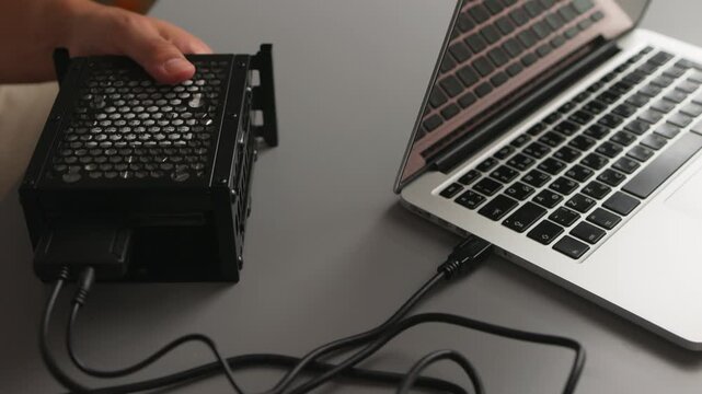 Hands connecting external hard drive enclosure to laptop with USB cable on tabletop, closeup of portable storage setup and hardware connection view from slight overhead angle, neutral soft lighting, t