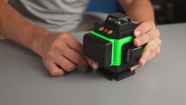 Hands adjusting compact cube laser level projecting green alignment lines on tabletop for precision measurement and calibration task in close-up view of tool and control knob as LED indicator glows.