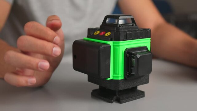 Hands adjusting compact cube laser level projecting green alignment lines on tabletop for precision measurement and calibration task in close-up view of tool and control knob as LED indicator glows.