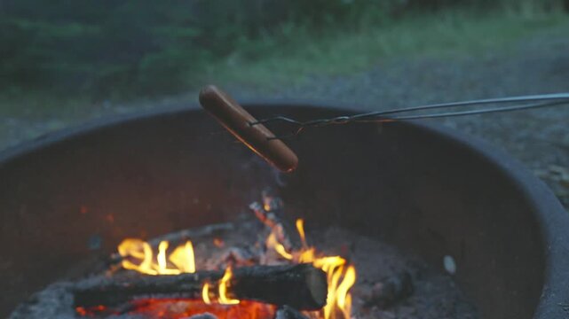 Roasting a hot dog on a metal skewer over a crackling campfire in a metal fire ring at a campsite