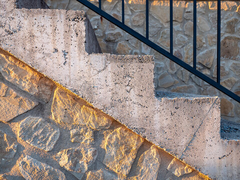 Close-up of outdoor concrete stairs with black metal railing and textured stone wall in warm evening sunlight. Architectural detail illustrating construction materials, geometry and exterior design.